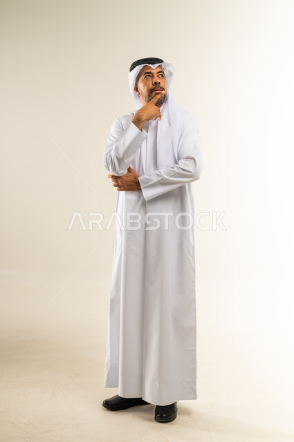 Anxiety and confusion in making the right decision, trying to find a solution to difficult problems, thinking deeply and absent-mindedness, portrait of a Kuwaiti Gulf Arab man wearing a traditional thobe and ghutra, putting his hand under his chin and looking somewhere far away with gestures of integration, full body, white background