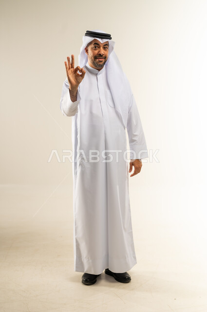 التعبير عن الإعجاب والتقدير، رفع اليد بعلامات الجودة والقبول، الامتياز والعلامة الكاملة، الشعور بالرضا والاستحسان، بورتريه لرجل عربي خليجي كويتي مبتسم يرتدي الثوب التقليدي والغتره ينظر إلى الكاميرا بإيماءات السعادة والسرور، الجسم بالطول الكامل، خلفية بيضاء