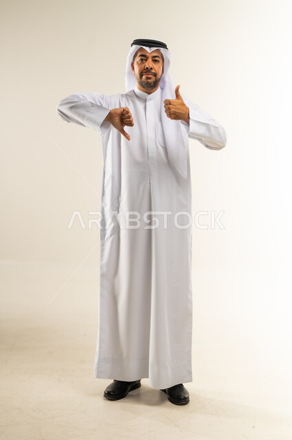 الحيرة في اتخاذ القرار الصحيح، الإشارة باصبعي الابهام إلى جهتين متعاكستين، علامة الجودة والسوء، الرضا وعدم الاستحسان، بورتريه لرجل عربي خليجي كويتي مبتسم يرتدي الثوب التقليدي والغتره يضع يده أسفل ذقنه وينظر إلى الكاميرا بجدية، الجسم بالطول الكامل، خلفية بيضاء