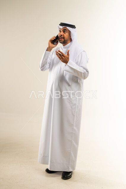 إجراء المكالمات الصوتية عبر الهاتف المحمول، التواصل مع الأهل والأصدقاء، دمج التقنيات الحديثة والتكنولوجيا في الحياة اليومية، بورتريه لرجل عربي خليجي كويتي مبتسم يرتدي الثوب التقليدي والغتره يجري مكالمة هاتفية بإيماءات الاندماج، الجسم بالطول الكامل، خلفية بيضاء