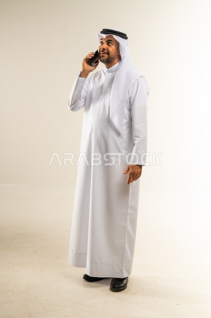 التواصل مع الأهل والأصدقاء، إجراء المكالمات الصوتية عبر الهاتف المحمول، دمج التقنيات الحديثة والتكنولوجيا في الحياة اليومية، بورتريه لرجل عربي خليجي كويتي مبتسم يرتدي الثوب التقليدي والغتره يجري مكالمة هاتفية بإيماءات الاندماج، الجسم بالطول الكامل، خلفية بيضاء