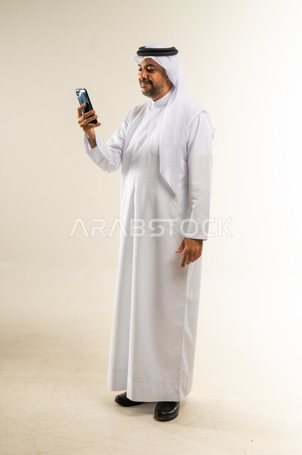 تصفح مواقع وتطبيقات الانترنت، النظر إلى الهاتف المحول بإيماءات الاندماج، دمج التقنيات والتكنولوجيا بالحياة اليومية، الدردشة والتحدث مع الأصدقاء، بورتريه لرجل عربي خليجي كويتي مبتسم يرتدي الثوب التقليدي والغتره يمسك الجوال بيده، الجسم بالطول الكامل، خلفية بيضاء