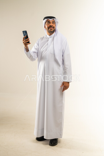 Looking somewhere far away with gestures of integration, integrating technology and techniques into daily life, chatting and talking with friends, browsing websites and applications, portrait of a smiling Kuwaiti Gulf Arab man wearing traditional thobe and ghutra holding mobile phone in hand, full body, white background