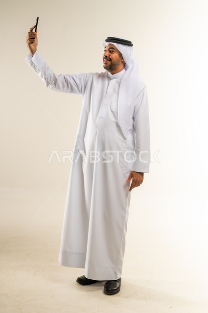 أخذ اللقطات الشخصية التذكارية، التقاط صور السيلفي باستخدام الهاتف المحمول، دمج التقنيات والتكنولوجيا الحديثة بالحياة اليومية، بورتريه لرجل عربي خليجي كويتي مبتسم يرتدي الثوب التقليدي والغتره يمسك الجوال بيده وينظر إليه بسعادة، الجسم بالطول الكامل، خلفية بيضاء