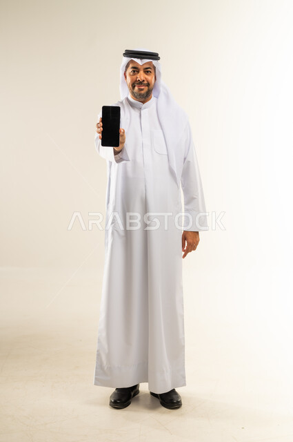 عرض شاشة هاتف محمول سوداء فارغة، دمج التقنيات والتكنولوجيا الحديثة بالحياة اليومية، الإعلانات التسويقية والعروض الترويجية، بورتريه لرجل عربي خليجي كويتي مبتسم يرتدي الثوب التقليدي والغتره يمسك الجوال بيده وينظر للكاميرا ببشاشة، الجسم بالطول الكامل، خلفية بيضاء