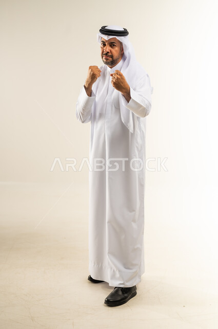 Professional boxing, hands closed and ready to attack and confront, trying to defend oneself, concept of strength and masculinity, portrait of a Kuwaiti Gulf Arab man wearing traditional thobe and ghutra looking at the camera with gestures of seriousness and anger, full body, white background