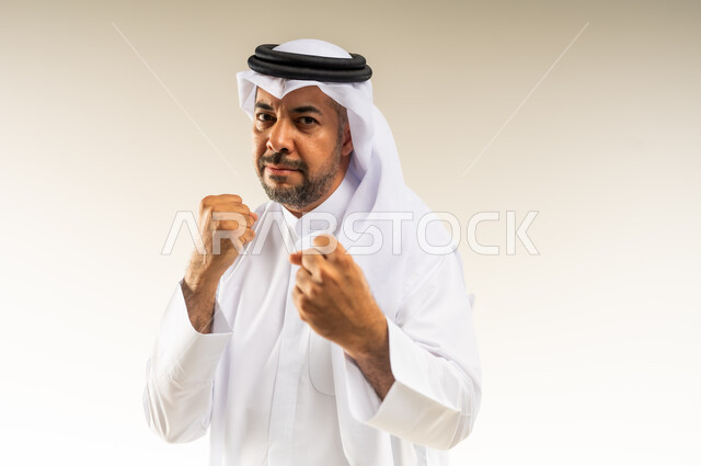 إغلاق اليدين والاستعداد للهجوم والمواجهة، احتراف رياضة الملاكمة، محاولة الدفاع عن النفس، مفهوم القوة والرجولة، بورتريه مقرب لرجل عربي خليجي كويتي  يرتدي الثوب التقليدي والغتره ينظر للكاميرا بإيماءات الجدية والغضب، خلفية بيضاء