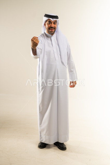 النجاح والفوز بشيء ما منتظر، الحماس والسعادة العارمة، الفرح الهستيري بتحقيق الأهداف والأحلام، بورتريه لرجل عربي خليجي كويتي مبتسم يرتدي الثوب التقليدي والغتره يرفع قبضته في الهواء وينظر إلى الكاميرا بإيماءات السرور، الجسم بالطول الكامل، خلفية بيضاء