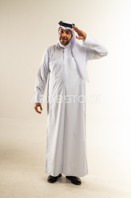 Elegance and interest in appearance, looking at the camera with gestures of cheerfulness and pleasure, the concept of handsomeness and self-confidence, portrait of a smiling Kuwaiti Gulf Arab man wearing traditional thobe adjusting and arranging his ghutra, full-length body, white background