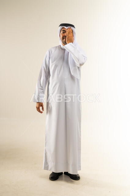 ضعف النظر وتشوش الرؤية، وضع اليد على العين وتغطيها، التهاب واحمرار العيون والحاجة لزيارة الطبيب، التعرض لأذية في الشبكية، بورتريه لرجل عربي خليجي كويتي يرتدي الثوب التقليدي والغترة يعاني من مشاكل صحية في عينه، الجسم بالطول الكامل، خلفية بيضاء