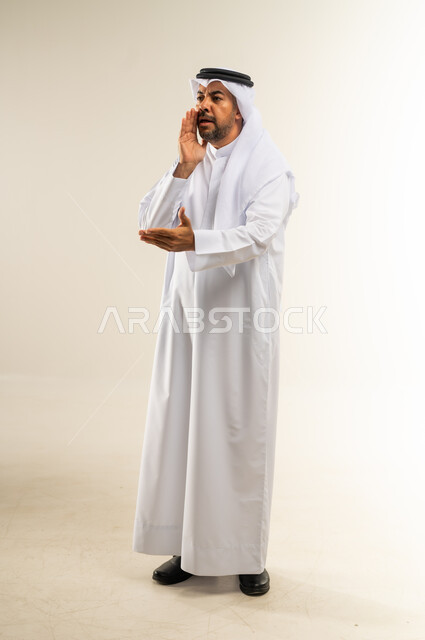 النداء بصوت مرتفع لأحد ما بمكان بعيد, وضع اليد بجانب الفم لزيادة حدة الأصوات، الصراخ والتكلم بنغمة عالية للسماع بشكل جيد، بورتريه لرجل عربي خليجي كويتي يرتدي الثوب التقليدي والغترة ينظر إلى مكان ما بإيماءات الاندماج والتركيز, الجسم بالطول الكامل، خلفية بيضاء