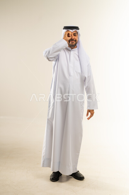 Trying to bring distant things closer, planning for future goals and ambitions, love of discovery, knowledge and adventure, focusing on something, portrait of a Kuwaiti Gulf Arab man wearing traditional thobe and ghutra, putting his hand on his eye in the shape of a lens with searching gestures, full body, white background