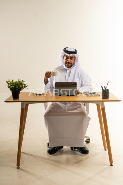 التلذذ بفنجان قهوة ساخن، تحقيق الأهداف بسرعة ومهارة باستخدام التقنيات الحديثة، التوظيف في المكاتب والشركات الكويتية، بورتريه لرجل عربي خليجي كويتي يرتدي الثوب التقليدي والغترة يجلس خلف المكتب يعمل على الجهاز اللوحي، الاحترافية والعمل الجاد، خلفية بيضاء