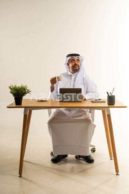 تحقيق الأهداف بسرعة ومهارة باستخدام التقنيات الحديثة، التلذذ بفنجان قهوة ساخن، التوظيف في المكاتب والشركات الكويتية، بورتريه لرجل عربي خليجي كويتي يرتدي الثوب التقليدي والغترة يجلس خلف المكتب يعمل على الجهاز اللوحي، الاحترافية والعمل الجاد، خلفية بيضاء