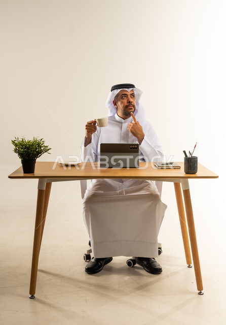 النظر إلى مكان ما بإيماءات الشرود والحيرة، الاستماع بتناول فنجان من القهوة الساخنة، التوظيف في المكاتب والشركات الكويتية، بورتريه لرجل عربي خليجي كويتي يرتدي الثوب التقليدي والغترة يجلس خلف المكتب بجانه نبته خضراء وأمامه الجهاز اللوحي، خلفية بيضاء