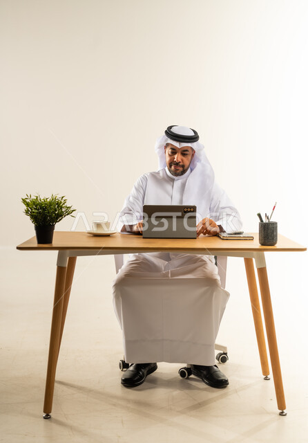 التركيز والعمل بجد وتفاني، استخدام الجهاز اللوحي في القطاعات المكتبية والإدارية، إنجاز المهام بسرعة واحترافية، التوظيف في المكاتب والشركات الكويتية، بورتريه لرجل عربي خليجي كويتي يرتدي الثوب التقليدي والغترة يجلس خلف المكتب ويشرف على سير الأعمال، خلفية بيضاء