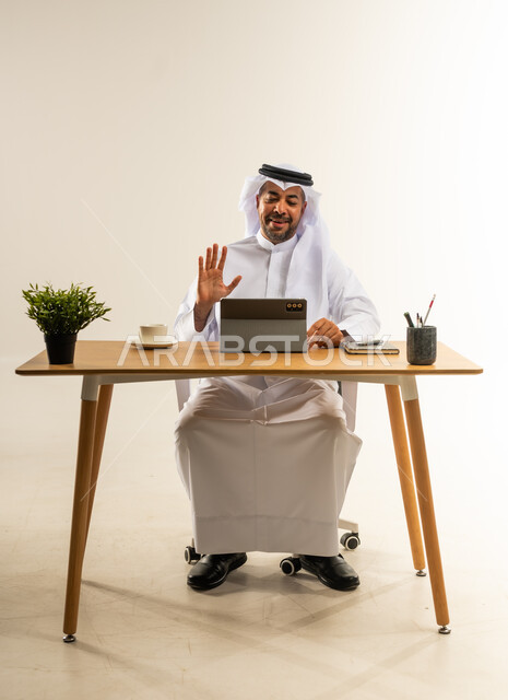 إجراء مكالمات الفيديو باستخدام الجهاز اللوحي في العمل، إنجاز المهام بسرعة واحترافية عالية، التوظيف في المكاتب والشركات الكويتية، بورتريه لرجل عربي خليجي كويتي يرتدي الثوب التقليدي والغترة يجلس خلف المكتب يجري اجتماع أونلاين عبر الانترنت، خلفية بيضاء