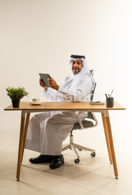 Utilizing modern technologies in administrative work, employment in Kuwaiti offices and companies, professionalism and speed in completing tasks, dedication, hard work and seriousness, portrait of a Kuwaiti Gulf Arab man wearing traditional thobe and ghutra sitting behind a desk and working on a tablet, white background