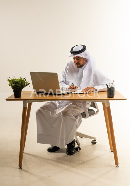 تسجيل وتدوين الملاحظات الهامة، دمج التقنيات الحديثة بالأعمال الإدارية والمكتبية، التركيز والتفاني العمل الجاد، التوظيف في المكاتب والشركات الكويتية، بورتريه لرجل عربي خليجي كويتي يرتدي الثوب التقليدي والغترة يجلس خلف المكتب وأمامه الحاسوب المحمول، خلفية بيضاء