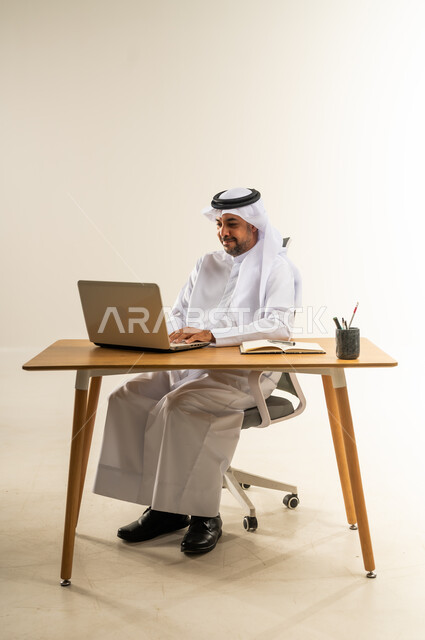 إنجاز المهام باستخدام الحاسوب المحمول، دمج التقنيات الحديثة بالأعمال الإدارية، التركيز والتفاني والعمل الجاد، التوظيف في المكاتب والشركات الكويتية، بورتريه لرجل عربي خليجي كويتي يرتدي الثوب التقليدي والغترة يجلس خلف طاولة المكتب ويعمل بجد، خلفية بيضاء