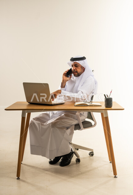استخدام التقنيات الحديثة والتكنولوجيا بالمجال الإداري، التوظيف في المكاتب والشركات الكويتية، الإجابة والرد على مكالمات العمل الهاتفية عبر الجوال، بورتريه لرجل عربي خليجي كويتي يرتدي الثوب التقليدي والغترة يجلس خلف المكتب وأمامه الحاسوب المحمول، خلفية بيضاء