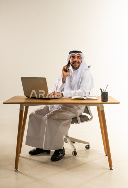 التوظيف في المكاتب الكويتية، الإجابة والرد على مكالمات العمل الهاتفية عبر الجوال، دمج التقنيات الحديثة والتكنولوجيا بالمجال الإداري والمكتبي، بورتريه لرجل عربي خليجي كويتي يرتدي الثوب التقليدي والغترة يجلس خلف المكتب وأمامه جهاز الحاسوب المحمول، خلفية بيضاء