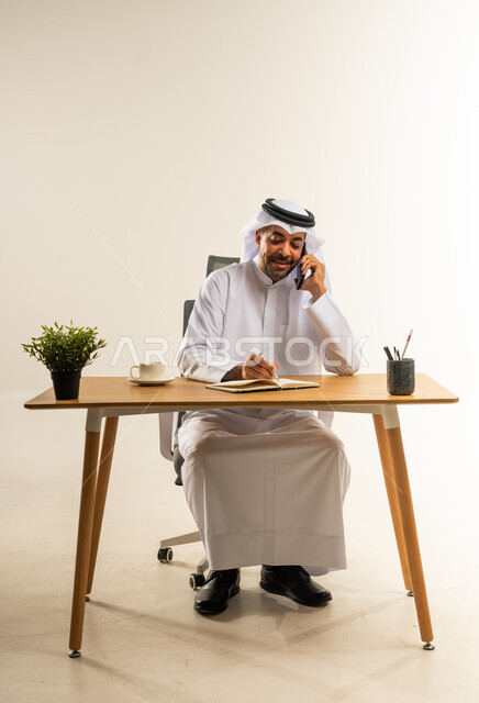 إجراء مكالمات العمل الهاتفية عبر الجوال، دمج التقنيات الحديثة بالمجال الإداري والمكتبي، التوظيف في المكاتب الكويتية، بورتريه لرجل عربي خليجي كويتي يرتدي الثوب التقليدي والغترة يجلس خلف المكتب بجانبه نبتة خضراء يقوم بالرد والإجابة على الاتصالات، خلفية بيضاء
