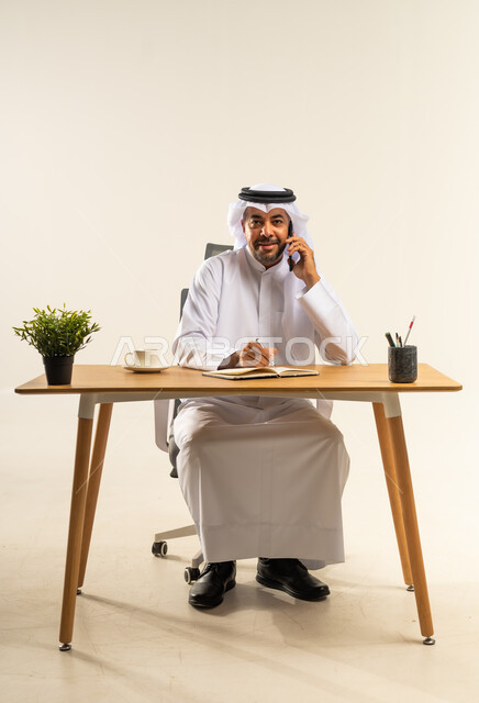 الرد والإجابة على اتصالات العمل، دمج التقنيات الحديثة والتكنولوجيا بالمجال الإداري والمكتبي، التوظيف في المكاتب الكويتية، بورتريه لرجل عربي خليجي كويتي يرتدي الثوب التقليدي والغترة يجلس خلف المكتب وبجانبه نبتة خضراء يجري مكالمة هاتفية عبر الجوال، خلفية بيضاء