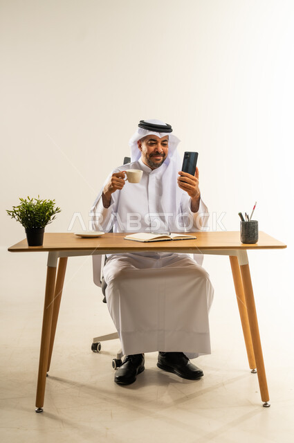 الاستمتاع بتناول فنجان قهوة ساخنة، دمج التقنيات الحديثة بالمجال الإداري والمكتبي، العمل في المكاتب والشركات الكويتية، بورتريه لرجل عربي خليجي كويتي يرتدي الثوب التقليدي والغترة يجلس خلف المكتب بجانبه نبتة خضراء ويمسك الهاتف المحمول، خلفية بيضاء