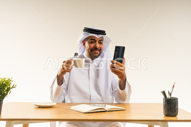 العمل في المكاتب والشركات الكويتية، الاستمتاع بتناول فنجان من القهوة الساخنة الغنية، دمج التقنيات الحديثة بالمجال الإداري، بورتريه مقرب لرجل عربي خليجي كويتي يرتدي الثوب التقليدي والغترة يجلس خلف المكتب بجانبه نبتة خضراء ويمسك الهاتف المحمول، خلفية بيضاء