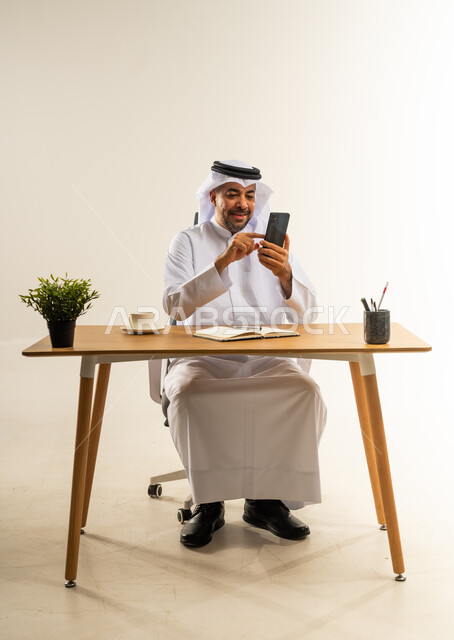 دمج التقنيات الحديثة بالمجال الإداري والمكتبي، العمل في المكاتب والشركات الكويتية، الاستمتاع بتناول فنجان قهوة ساخنة، بورتريه لرجل عربي خليجي كويتي يرتدي الثوب التقليدي والغترة يجلس خلف المكتب بجانبه نبتة خضراء ويمسك الهاتف المحمول، خلفية بيضاء