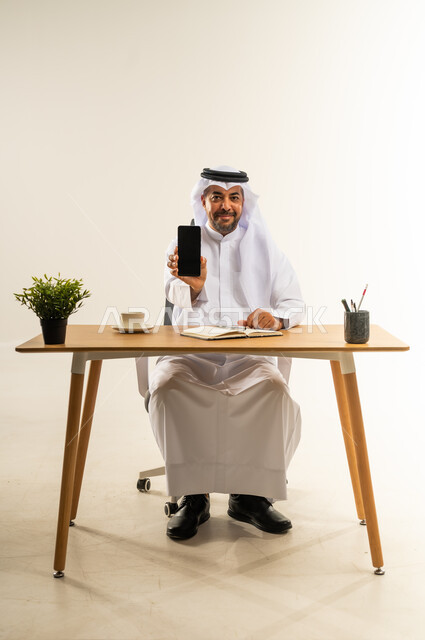 عرض شاشة جوال سوداء فارغة، دمج التقنيات الحديثة بالمجال الإداري والمكتبي، العمل في المكاتب والشركات الكويتية، بورتريه لرجل عربي خليجي كويتي يرتدي الثوب التقليدي والغترة يجلس خلف المكتب وبجانبه نبتة خضراء وفنجان من القهوة الساخنة، خلفية بيضاء