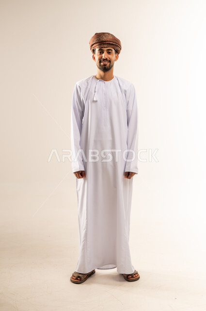 الاهتمام بالأناقة والمظهر الخارجي، مفهوم القوة والرجولة والشياكة، بورتريه لرجل عربي خليجي عماني مبتسم يرتدي الدشداشه والعمامة ينظر الى مكان ما في الاعلى بتعابير الفرح والسرور ، ايماءات الثقة بالنفس، الجسم الكامل بالطول، خلفية بيضاء