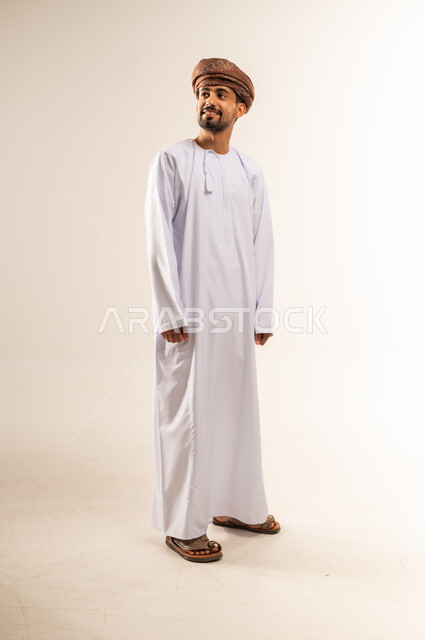 Concern for elegance and outward appearance, the concept of strength, masculinity and chic, portrait of a smiling Omani Gulf Arab man wearing a dishdasha and turban looking somewhere above with expressions of joy and happiness, gestures of self-confidence, full body length, white background