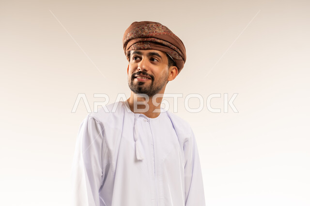 مفهوم القوة والرجولة والشياكة، الاهتمام بالأناقة والمظهر الخارجي، بورتريه لرجل عربي خليجي عماني مبتسم يرتدي الدشداشه والعمامة ينظر الى مكان ما في الاعلى بتعابير الفرح والسرور ، ايماءات الثقة بالنفس، خلفية بيضاء