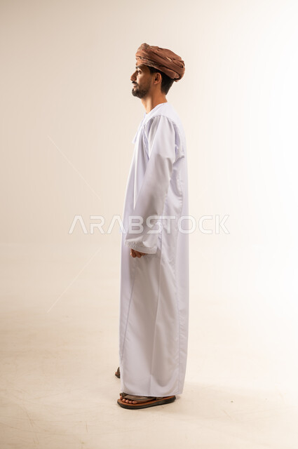 مفهوم القوة والرجولة، الاهتمام بالأناقة والمظهر الخارجي، بورتريه من الجانب لرجل عربي خليجي عماني يرتدي الدشداشه والعمامة يقوم بالنظر الى مكان ما جانبا، ايماءات الثقة بالنفس، الجسم الكامل بالطول، خلفية بيضاء
