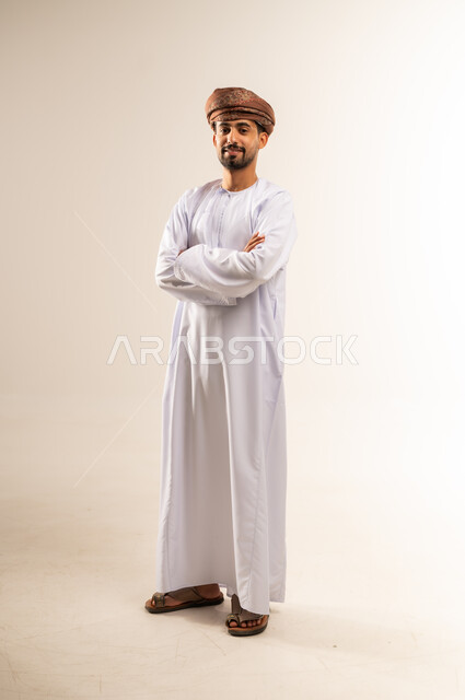 الاهتمام بالأناقة والمظهر الخارجي، مفهوم القوة والرجولة والشياكة، بورتريه لرجل عربي خليجي عماني يرتدي الدشداشه والعمامة يقف بإيماءات ايدي متقاطعة ينظر الى الكاميرا بتعابير الفرح والسرور والثقة بالنفس، الجسم الكامل بالطول، خلفية بيضاء