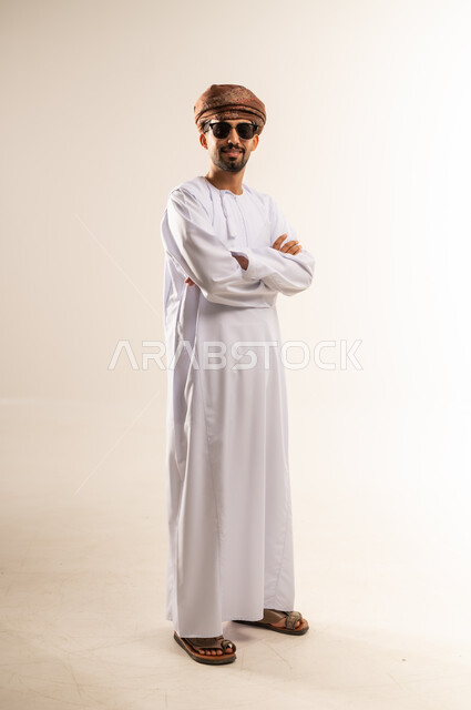 Concern for elegance and outward appearance, the concept of strength, masculinity and chic, portrait of an Arab Gulf Omani man wearing a dishdasha, turban and sunglasses, standing with crossed hands gestures, looking at the camera with expressions of joy, happiness and self-confidence, full body length, white background