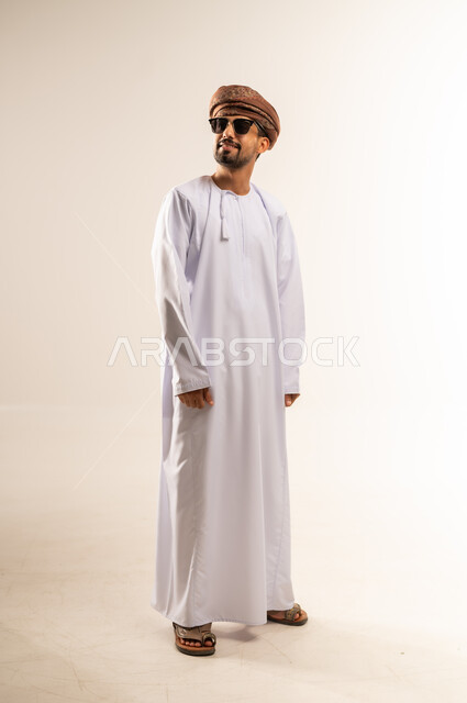 مفهوم القوة والرجولة والوسامة، الاهتمام بالأناقة والمظهر الخارجي، بورتريه لرجل عربي خليجي عماني مبتسم يرتدي الدشداشه والعمامة والنظارات الشمسية ينظر الى مكان ما بتعابير الفرح والسرور، ايماءات الثقة بالنفس، الجسم الكامل بالطول، خلفية بيضاء