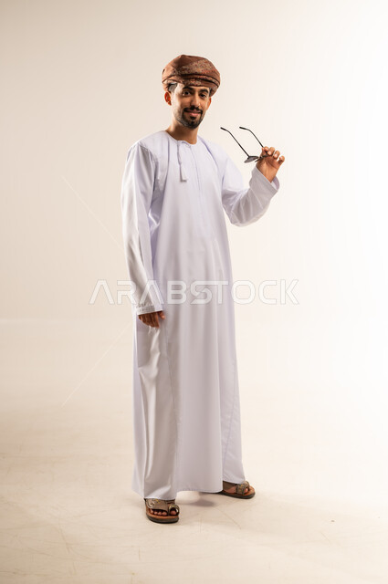 Concern for elegance and appearance, concept of strength and masculinity, portrait of a smiling Omani Gulf Arab man wearing a dishdasha and turban, holding sunglasses in his hand and looking at the camera with expressions of joy and happiness, gestures of self-confidence, full body length, white background