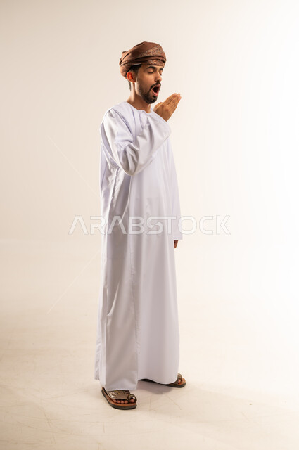 الشعور بالتعب والحاجة الى النوم والراحة, الإحساس بالكسل والخمول و الإرهاق، بورتريه لشاب عربي خليجي عماني يرتدي الدشداشة و العمامة يضع يده على فمه بإيماءات التثاؤب والنعاس، صورة الجسم بالطول الكامل, خلفية بيضاء