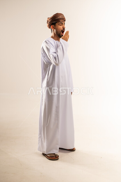 الشعور بالتعب والحاجة الى النوم والراحة, الإحساس بالكسل والخمول و الإرهاق، بورتريه لشاب عربي خليجي عماني يرتدي الدشداشة و العمامة يضع يده على فمه بإيماءات التثاؤب والنعاس، صورة الجسم بالطول الكامل, خلفية بيضاء