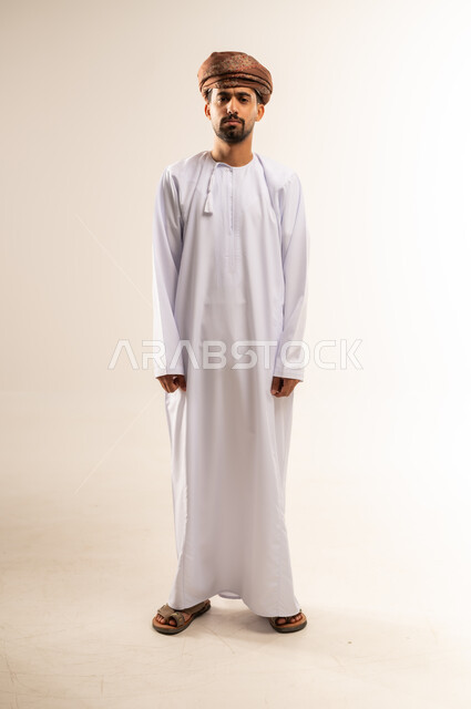 الاهتمام بالأناقة والمظهر الخارجي، مفهوم القوة والرجولة، بورتريه لرجل عربي خليجي عماني يرتدي الدشداشه والعمامة ينظر الى الكاميرا بتعابير الحزم والجدية، ايماءات الثقة بالنفس، الجسم الكامل بالطول، خلفية بيضاء