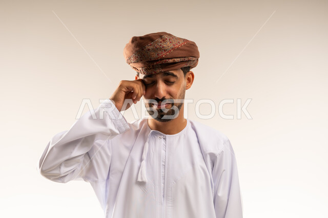 مفهوم تعديل الرؤية وضعف البصر، مشاكل صحية في العيون، الاحساس بألم في العينين، بورتريه لشاب عربي خليجي عماني يرتدي الدشداشة و العمامة يضع يديه على عينيه بتعابير التعب الشديد والصداع،  الشعور بالارهاق وعدم الراحة, خلفية بيضاء