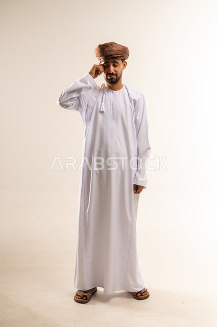 مفهوم تعديل الرؤية وضعف البصر، مشاكل صحية في العيون، الاحساس بألم في العينين، بورتريه لشاب عربي خليجي عماني يرتدي الدشداشة و العمامة يضع يديه على عينيه بتعابير التعب الشديد والصداع،  الشعور بالارهاق وعدم الراحة، صورة الجسم بالطول الكامل, خلفية بيضاء