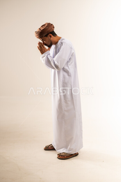 الطلب بترجي وتذلل وخضوع، مفهوم الشكر والامتنان، الوقوف بإيماءات الدعاء والتوسل والاسترحام، بورتريه من الجانب لرجل عربي خليجي عماني يرتدي الدشداشة والعمامة ينحني ويرفع يديه بخشوع واستعطاف، طلب العون والرغبة في تحقيق الأمنيات، الجسم بالطول الكامل، خلفية بيضاء