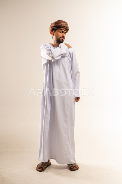 الأوجاع والآلام العصبية والعضلية، طلب المساعده والحاجه للعلاج، التهاب العضلات والمفاصل، بورتريه لشاب عربي خليجي عماني يرتدي الدشداشة و العمامة يضع يده على كتفه بايماءات الألم والتعب والأرهاق، حالة اسعافية طارئة، صورة الجسم الطول الكامل, خلفية بيضاء