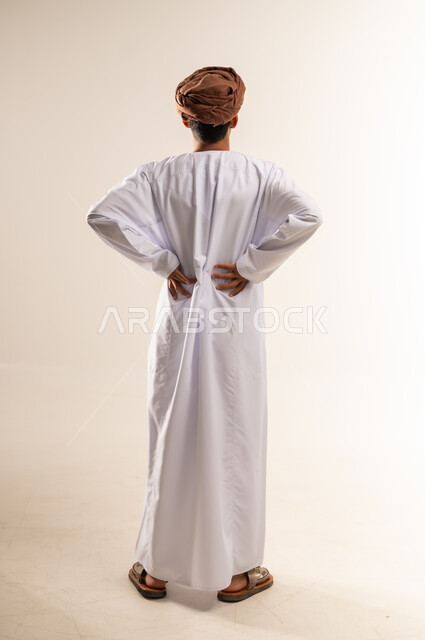 الحاجه للراحه من التعب والإرهاق , الاحساس بألم في العمود الفقري , بورتريه من الخلف  لرجل عربي خليجي عماني يرتدي الدشداشة والعمامة يضع يده اسفل ظهره بتعابير الشعور بالوجع, طلب المساعده للذهاب الى الطبيب ,صورة الجسم بالكامل، خلفية بيضاء