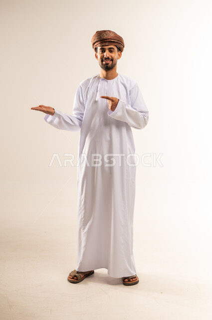 النظر إلى الكاميرا بإيماءات الفرح والسرور، بورتريه لشاب عربي خليجي عماني مبتسم يرتدي الدشداشة والعمامة يقف ويشير بإصبعيه السبابة جانبا  إلى شيء محدد بتعابير هذا المكان هنا، الجسم الكامل بالطول، خلفية بيضاء .