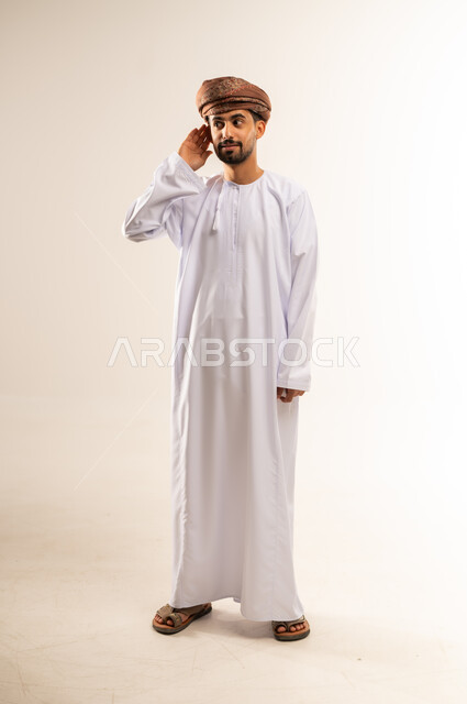 الانصات وحسن الاستماع، التفاجؤ من شيء ما، بورتريه لشاب عربي خليجي عماني مبتسم يرتدي الدشداشة و العمامة يضع يده خلف أذنه بإيماءات التنصت والتجسس، التركيز على الكلام وسماع الاسرار، الجسم الكامل بالطول، خلفية بيضاء